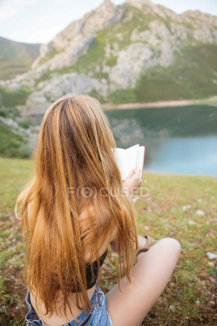 Visão traseira do livro de leitura de menina loira na costa do lago de montanha — Fotografia de Stock