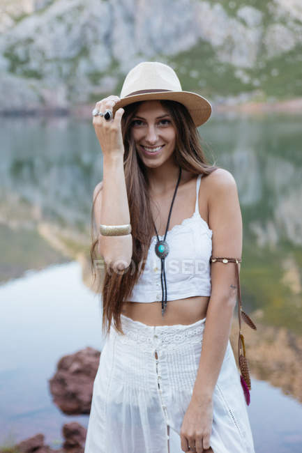 Brünettes Mädchen mit weißem Kleid, Hut und Blick in die Kamera am Bergsee — Stockfoto