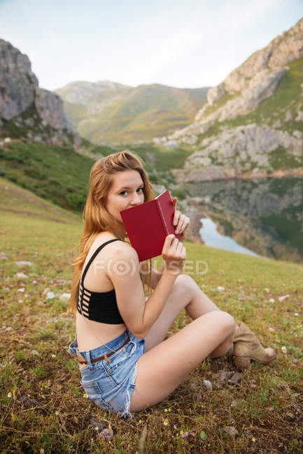 Young girl sitting on grass with book with mountains on background. — Stock Photo