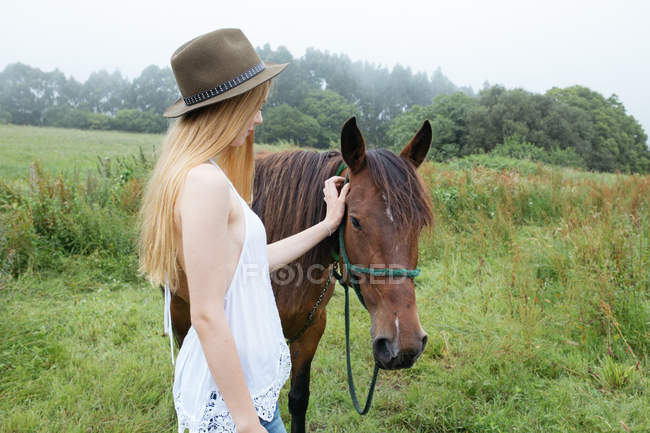 Девушка в шляпе гладит коричневую лошадь — стоковое фото