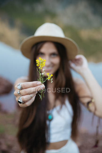 Portrait of brunette woman in hat showing yellow flower at camera — Stock Photo