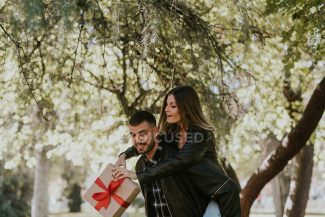 Cheerful man giving piggy back to girlfriend with gift at park — Stock Photo