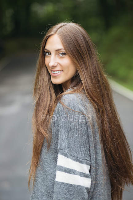 Retrato de menina morena vestindo camisola cinza em pé na estrada e olhando por cima do ombro para a câmera — Fotografia de Stock