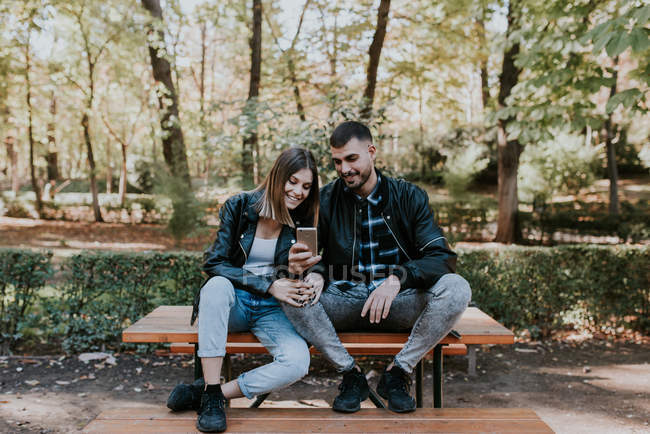 Couple souriant assis sur la table du parc et navigateur smartphone — Photo de stock