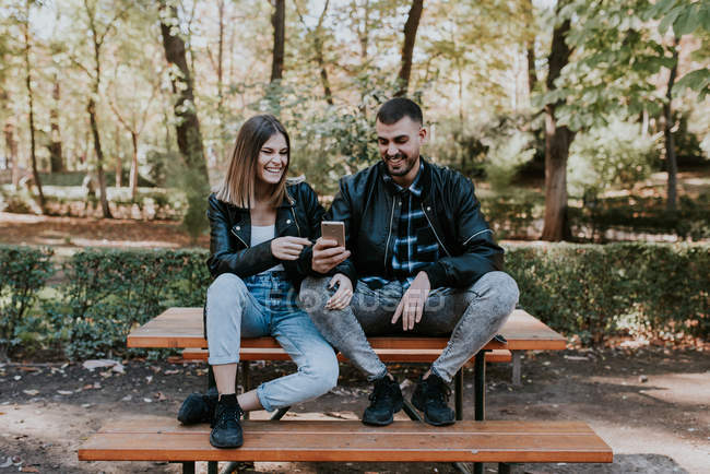 Couple souriant assis sur la table du parc et navigateur smartphone — Photo de stock