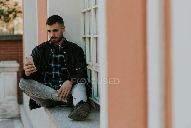 Молодой человек сидит на подоконнике и просматривает смартфон — стоковое фото
