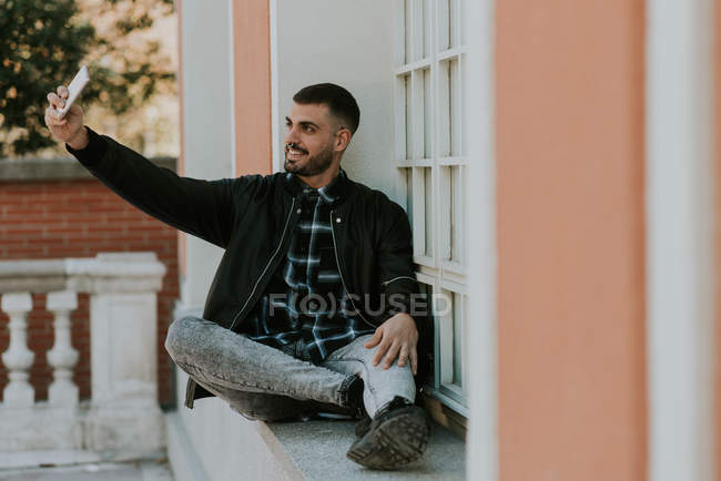 Retrato de hombre con chaqueta negra sentado en el alféizar de la ventana tomando selfie con smartphone - foto de stock