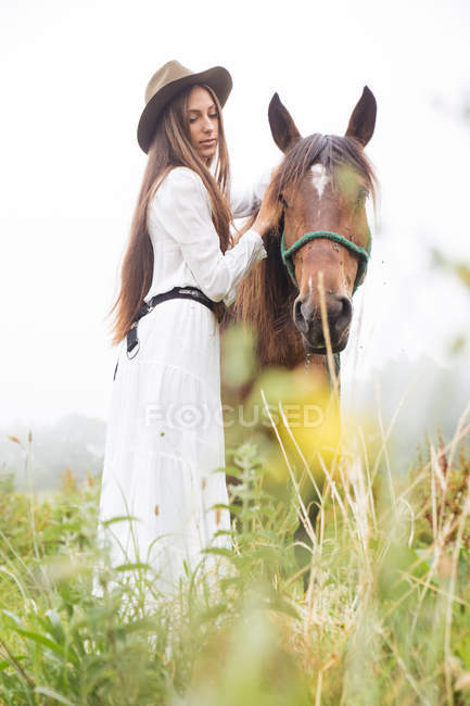 Вид збоку молодої брюнетки в білому платті обіймає коричневого коня в полі — стокове фото
