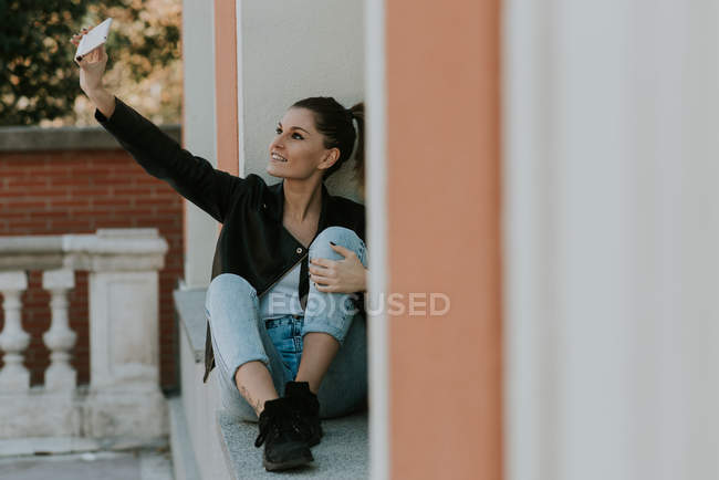 Retrato de una chica tomando selfie mientras está sentada en el alféizar de la ventana - foto de stock
