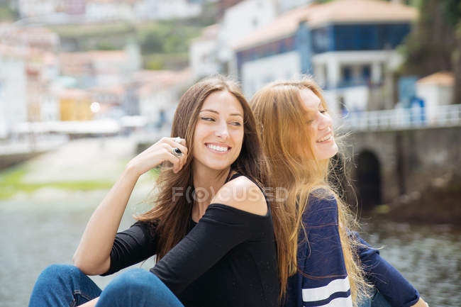 Portrait of cheerful best friends sitting back to back in street — Stock Photo