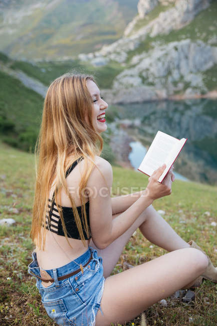 Сміється жінка з довгим волоссям тримає книгу, сидячи на траві біля гірського озера — стокове фото