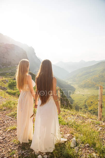 Back view of two young  girlfriends watching beautiful mountain landscape in sunlight. — Stock Photo