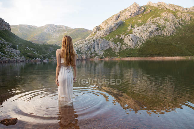 Rear view of blonde girl wearing white dress standing in mountain lake — Stock Photo