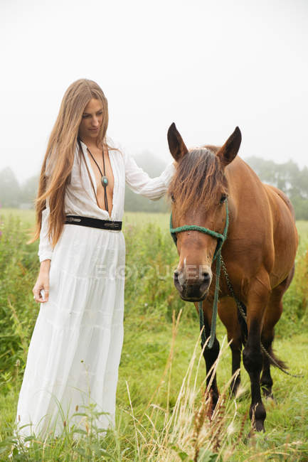 Menina de vestido branco em pé perto de cavalo marrom no campo — Fotografia de Stock