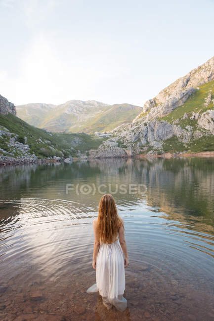 Vista trasera de la mujer de pie en el lago de montaña en vestido blanco - foto de stock