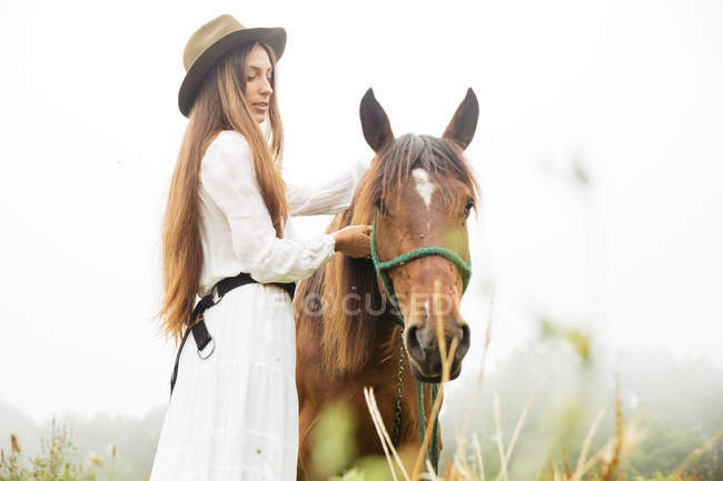 Vista laterale della giovane bruna in abito bianco abbraccia cavallo marrone in campo — Foto stock