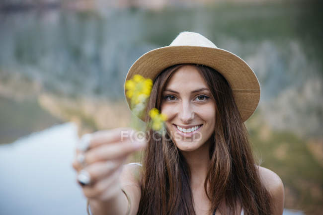 Portrait of brunette woman in hat showing yellow flower at camera against of lake on background — Stock Photo