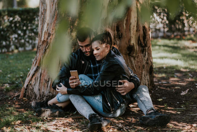 Paar umarmt sich auf dem Boden am Baum sitzend und surft mit dem Smartphone — Stockfoto
