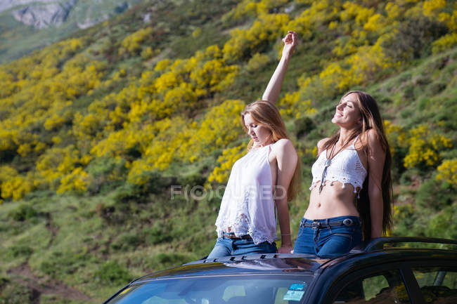 Dos novias felices sentadas en la escotilla del coche y disfrutando de la luz del sol en las montañas - foto de stock