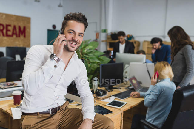 Retrato del hombre sonriente sentado en la mesa y hablando por teléfono sobre los trabajadores de oficina en segundo plano - foto de stock