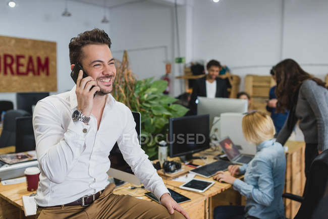 Retrato del hombre sonriente sentado en la mesa y hablando por teléfono en la oficina - foto de stock