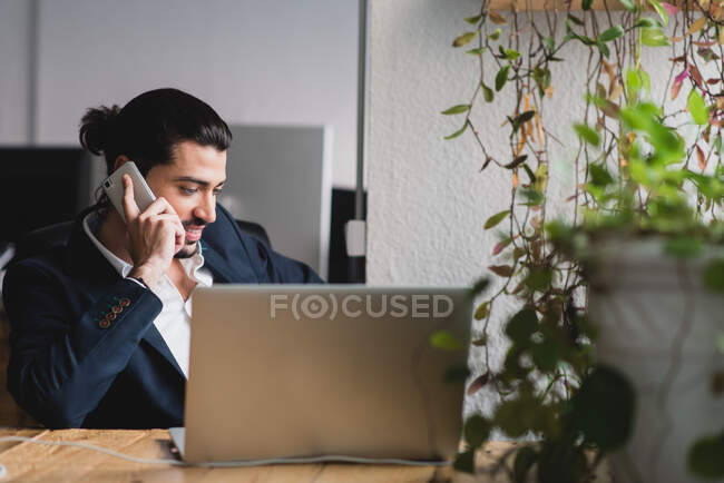 Confident businessman sitting at his laptop and talking phone. Horizontal indoors shot. — Stock Photo