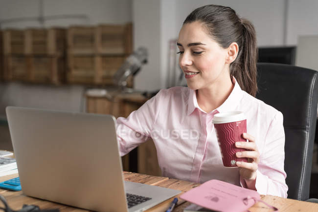 Улыбающаяся деловая женщина, держащая кофе и просматривающая ноутбук в офисе — стоковое фото