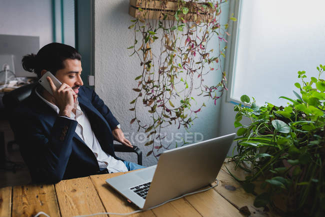 Retrato del hombre de negocios sentado en el lugar de trabajo con el ordenador portátil y tener conversación telefónica - foto de stock