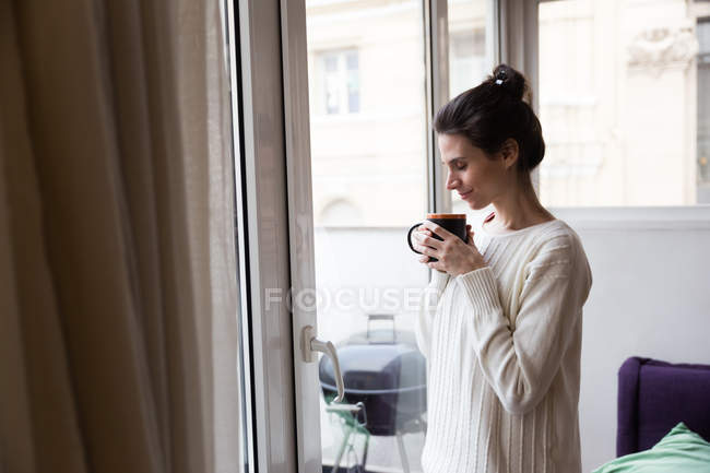 Vista laterale della donna che posa vicino alla finestra e coppa odorosa in mano con gli occhi chiusi — Foto stock