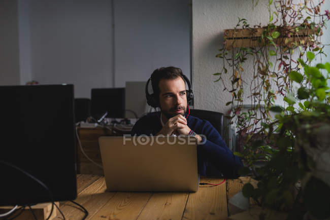 Retrato de homem em fones de ouvido sentado à mesa com laptop e olhando para longe — Fotografia de Stock