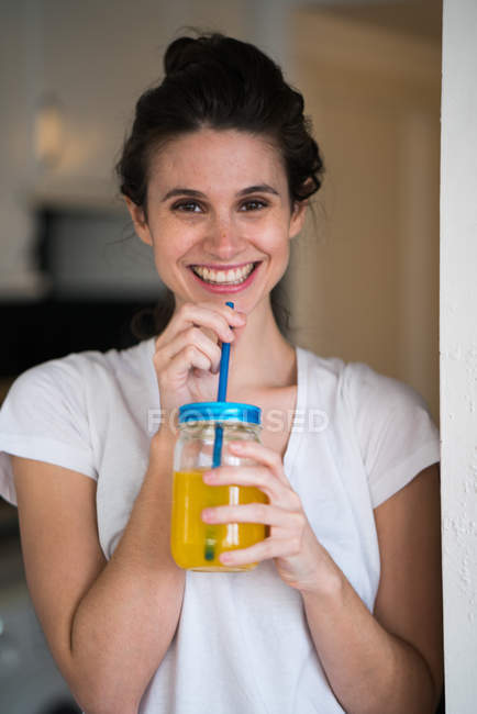 Portrait of smiling brunette woman drinking orange juice from glass jar and looking at camera — Stock Photo