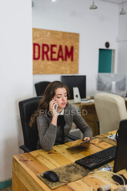 Retrato de una mujer joven sosteniendo una tableta y hablando por teléfono en el lugar de trabajo - foto de stock