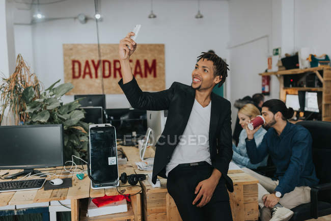 Portrait of smiling man sitting on table and making selfie at office — Stock Photo