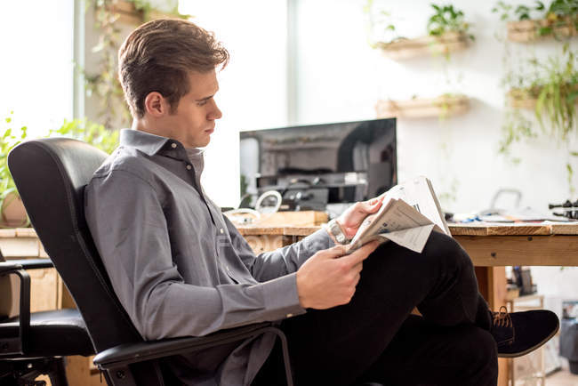 Young businessman reading newspaper while sitting in office chair. — Stock Photo