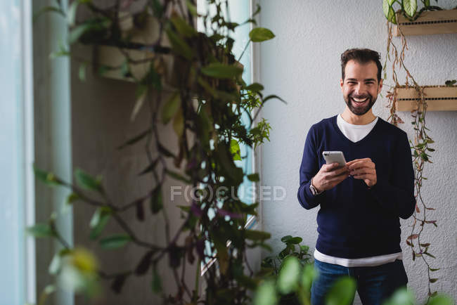 Портрет улыбающегося мужчины со смартфоном, стоящего у окна в офисе и смотрящего в камеру — стоковое фото
