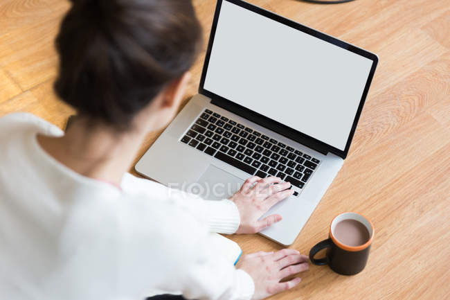 Через плечо вид женщины, пьющей какао и просматривающей ноутбук на полу — стоковое фото