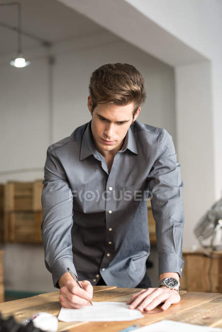 Молодой бизнесмен, стоящий за столом и подписывающий бумаги — стоковое фото