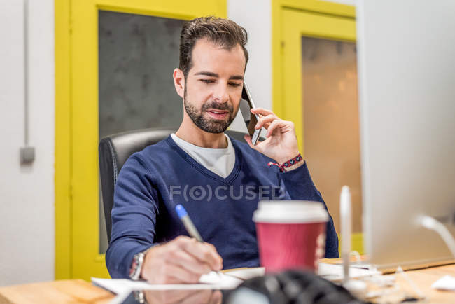 Thoughtful man writing on paper while talking on phone at office workplace — Stock Photo