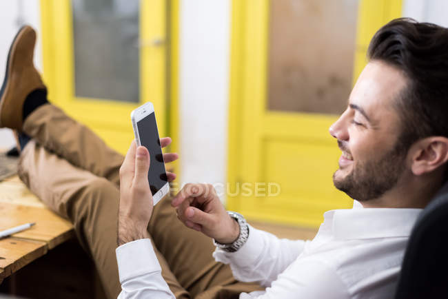 Side view of man sitting in chair with legs on table and browsing smartphone — Stock Photo