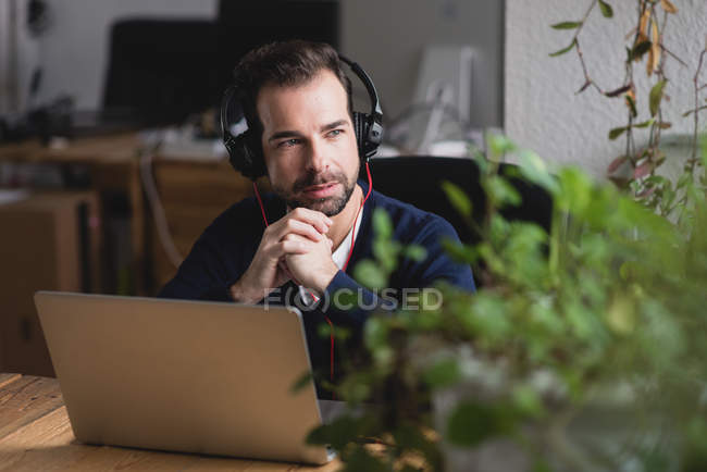 Retrato de hombre en auriculares sentado en la mesa con el ordenador portátil mirando a la ventana - foto de stock
