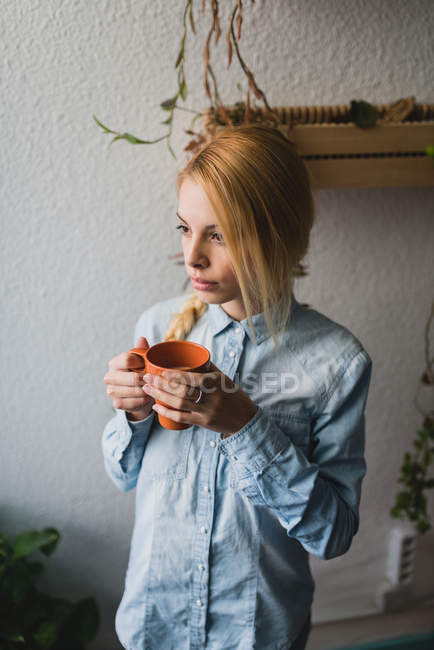 Portrait of blonde girl holding cup of tea and looking at window — Stock Photo