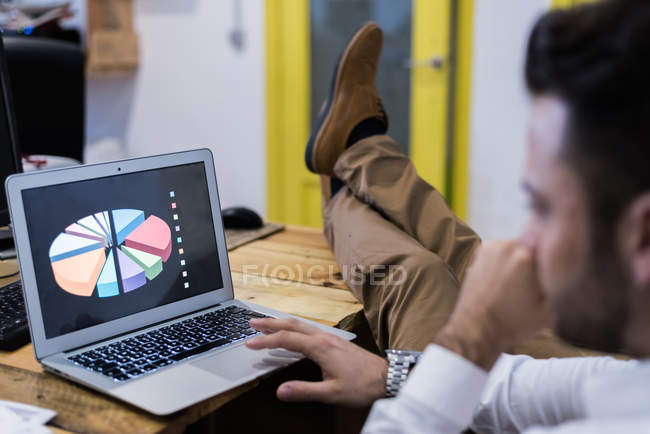 Seitenansicht eines Mannes mit Beinen auf dem Tisch beim Betrachten des Diagramms auf dem Laptop. — Stockfoto