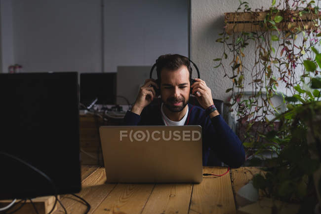 Retrato de homem sentado à mesa ajustando fones de ouvido e olhando para baixo laptop — Fotografia de Stock