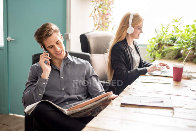 Чоловік розмовляє телефоном і дивиться на газету, коли жінка працює на ноутбуці . — стокове фото