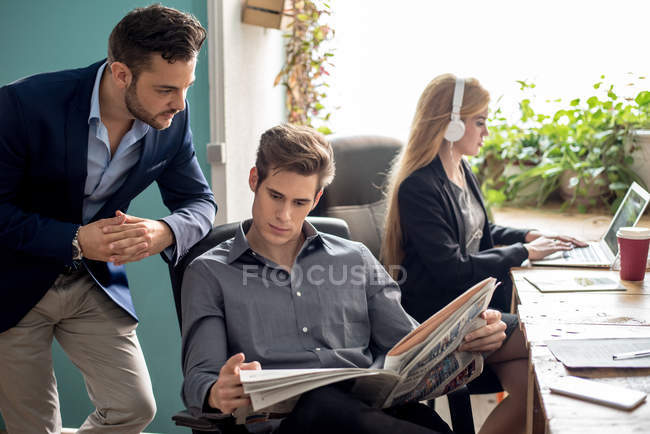 Dos colegas mirando el periódico mientras la mujer trabaja - foto de stock