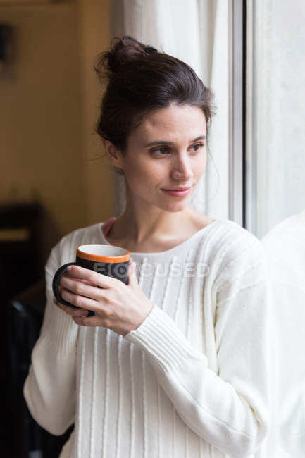 Portrait of young brunette girl posing with cup near window and looking aside — Stock Photo