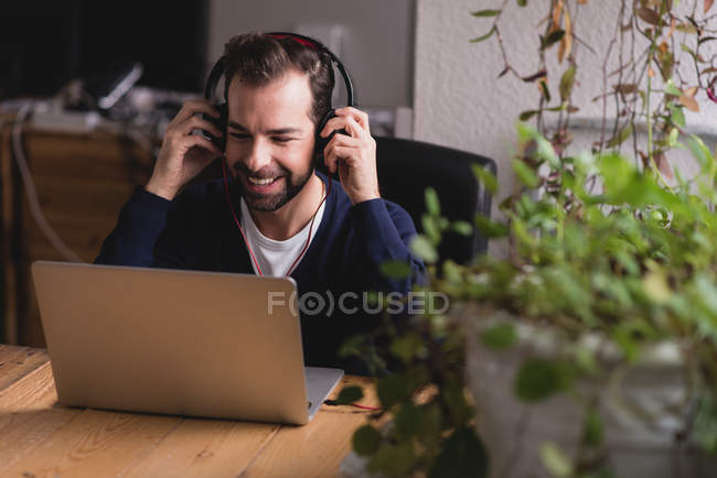 Retrato de homem sentado à mesa ajustando fones de ouvido e olhando para baixo laptop — Fotografia de Stock