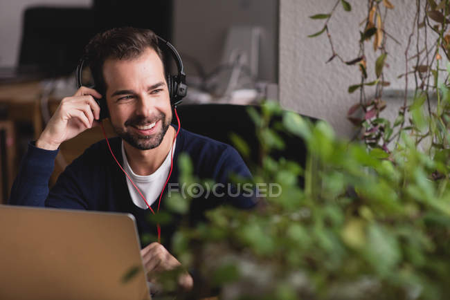 Retrato del hombre sonriente sentado en la mesa con el ordenador portátil ajustando los auriculares y mirando a la ventana - foto de stock