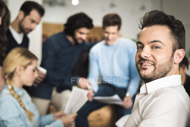 Веселий чоловік посміхається на камеру над групою людей, що працюють в офісі — стокове фото
