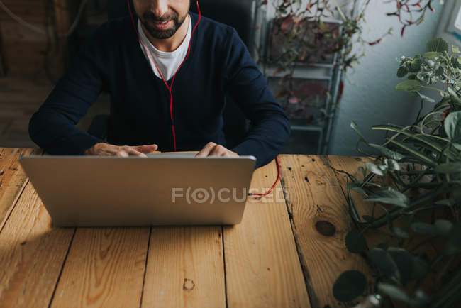 Abgeschnittenes Bild eines Mannes mit Kopfhörern, der am Tisch sitzt und eine Arbeitsfläche benutzt — Stockfoto